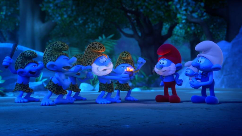 Papa Schlumpf steht mit Handy und Baby vor einer Gruppe von Schlümpfen mit Leoparden-Mützen und Lendenschürzen. Einer von ihnen hält ein Gefäß mit einer leuchtenden Flamme.