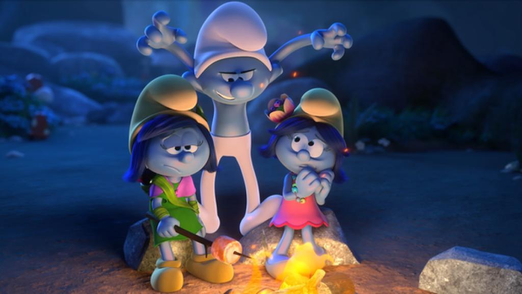 Jokey steht hinter zwei Schlumpfinnen, die am Lagerfeuer sitzen. Er reißt die Arme in die Höhe wie ein Monster und hat einen grimmigen Gesichtsausdruck. Schlumpfin Sturm schaut gelangweilt, Schlumpfin Blüte schaut ängstlich.