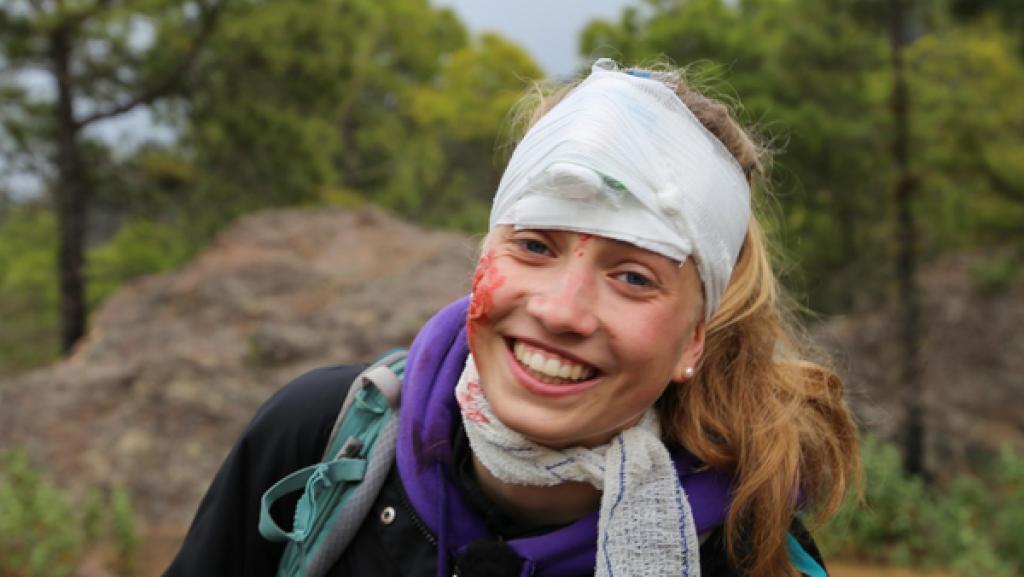 Ella hat Kunstblut im Gesicht und einen Kopfverband. Sie spielt ein Unfallopfer und lächelt fröhlich.