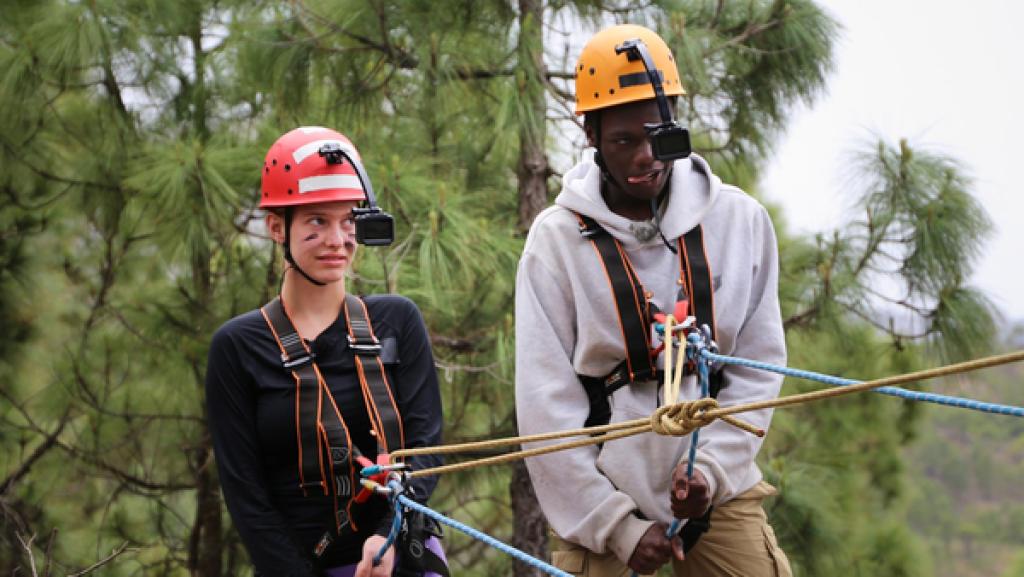 Tania und Isaak haben Helme auf und hängen in der Seilsicherung an einem Felsabhang.