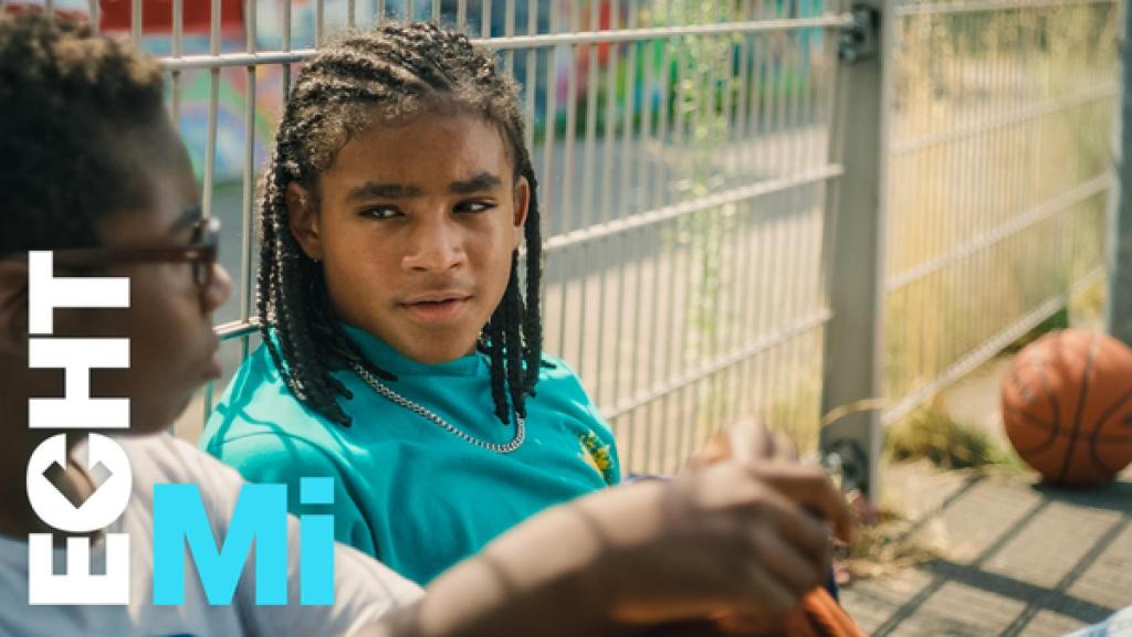 Elias (rechts) und Finn (links) unterhalten sich auf dem Basketballplatz. Beide sitzen auf dem Boden und lehnen sich an einen Zaun.