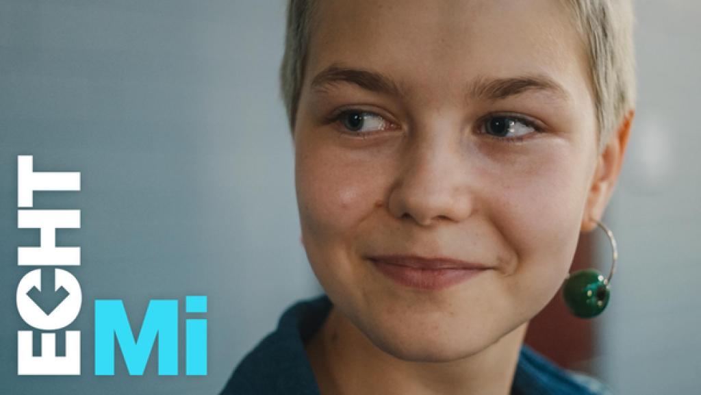 Jo sieht zur Seite und lächelt. Im linken Ohr trägt sie eine Kreole mit einer großen, grünen Perle.