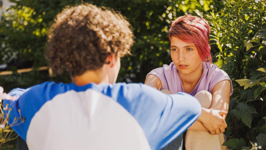 Ein jugendliches Mädchen und ein jungendlicher Junge sitzen draußen und schauen sich an