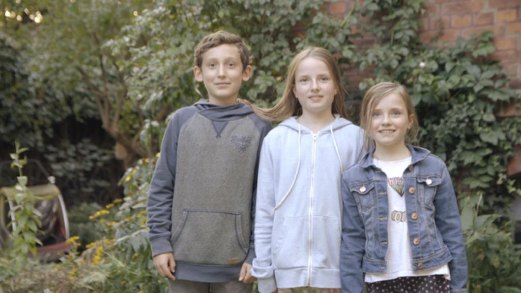 Zwei Mädchen und ein Junge stehen draußen nebeneinander vor einer bewachsenen Mauer.