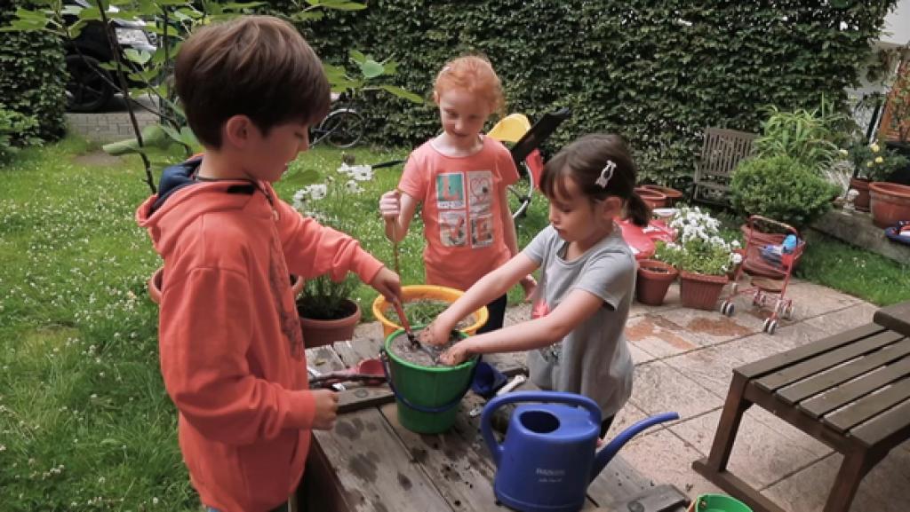 Zwei Mädchen und ein Junge stehen im Garten und mischen Schlamm mit Gräsern in Eimern.