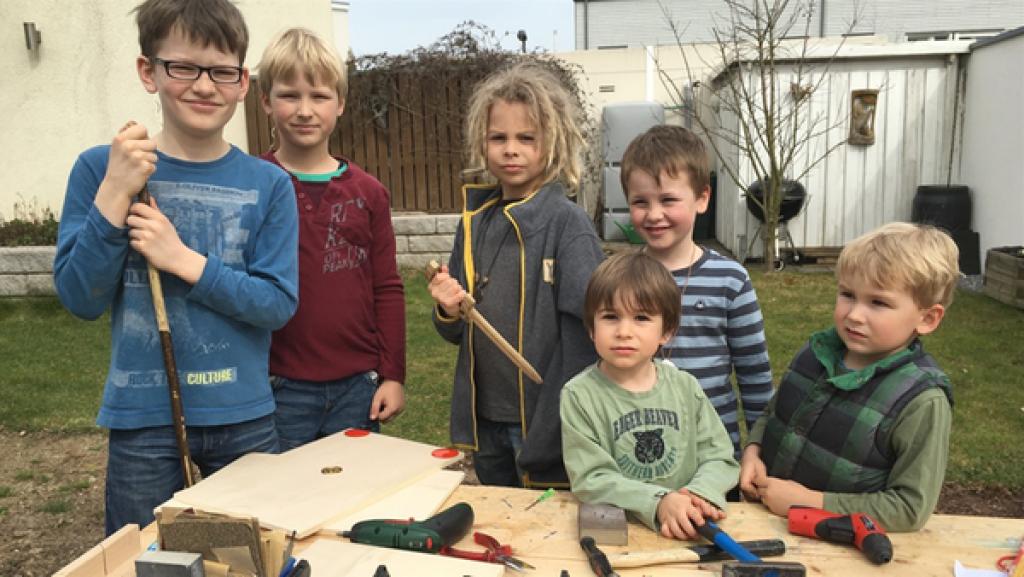 Sechs Kinder stehen an einer Werkbank. Vor ihnen liegt Holz, Schrauben und Werkzeug.