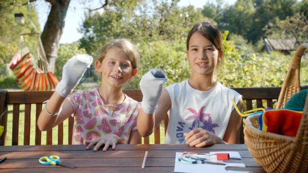 Zwei Mädchen sitzen draußen an einem Tisch und halten in einer Hand ihre gebastelten Socken.