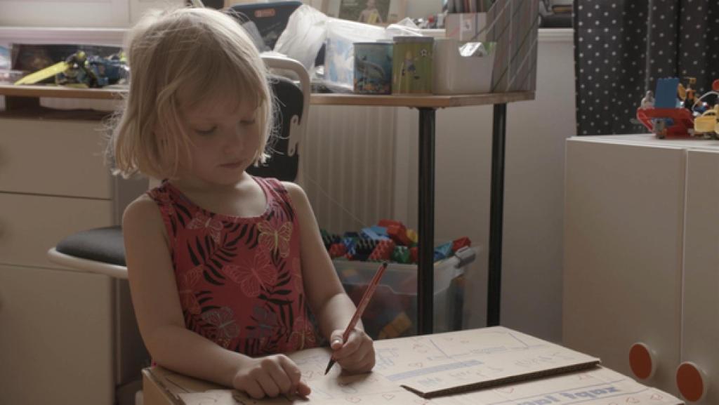 Ein Mädchen kniet auf dem Boden und hält einen roten Stift in der Hand. Sie malt auf einen Karton.