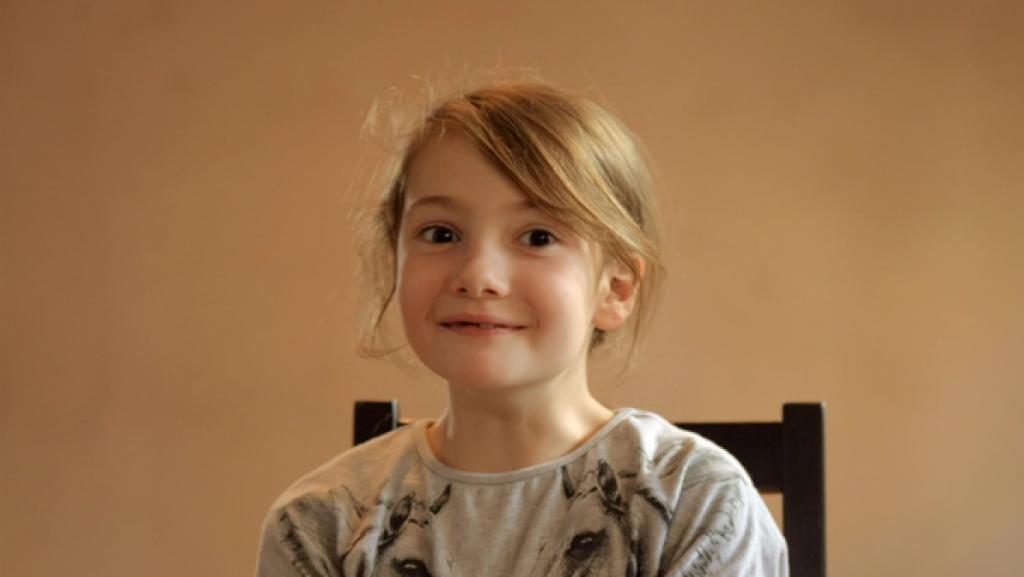 Ein etwa siebenjähriges Kind sitzt auf einem Stuhl und lächelt in die Kamera.