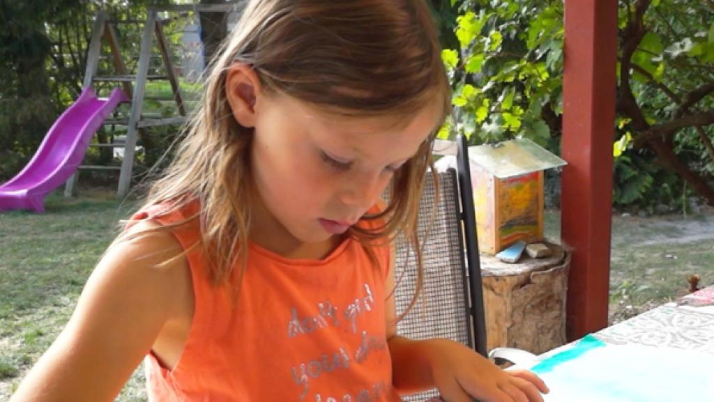 Ein Grundschulkind sitzt an einem Tisch im Garten und malt mit Fingermalfarben auf weißes Papier.