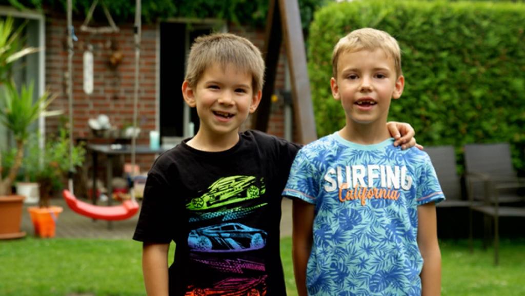 Zwei Jungs stehen draußen im Garten. Der linke Junge legt seinen Arm um den anderen. Beide lachen.