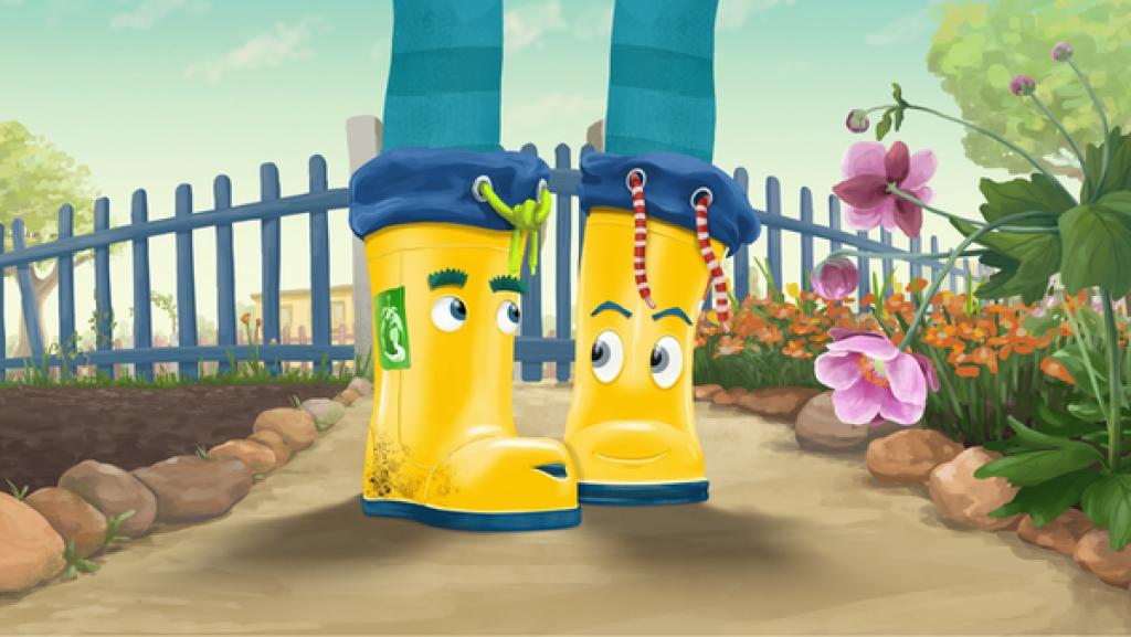 Der Gummistiefel Torf und der Gummistiefel Buddel stehen in einem Garten. Rechts von Ihnen blühen rosa und rote Blüten. Links von ihnen steht ein Beet leer.