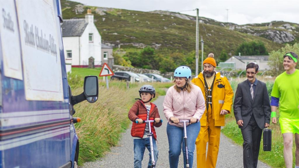 Sam schaut sich zusammen mit einem Mädchen und drei Männern einen mobilen Verkaufswagen an, der auf einer kleinen Straße steht.