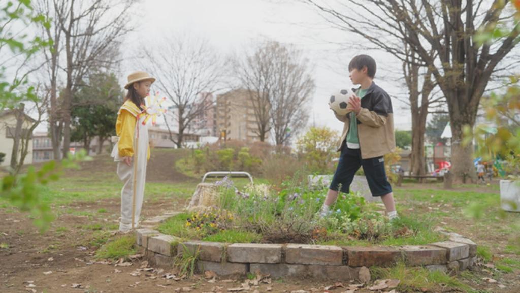 Ein Junge mit einem Fußball in den Händen trampelt über ein Blumenbeet. Daneben steht ein Mädchen, das ihn vorwurfsvoll anschaut.