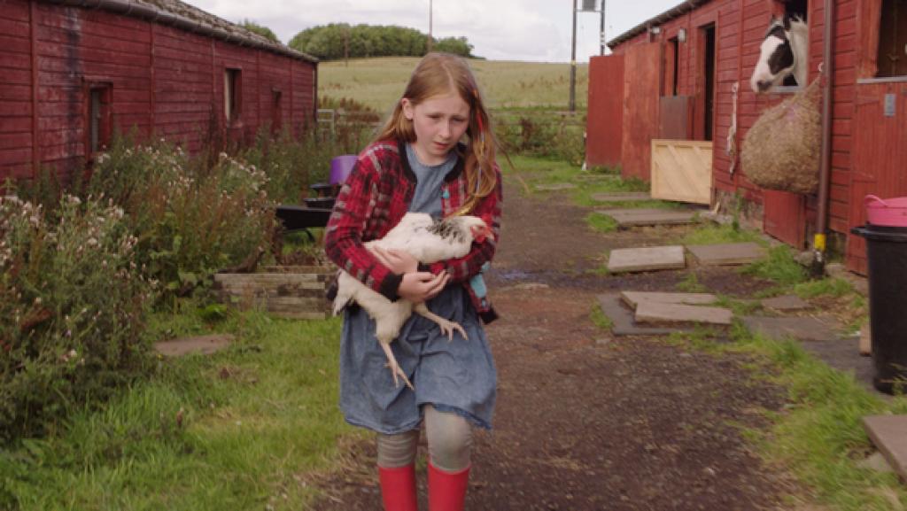 Rechte: SR/BBC Alba/Schottland/EBU 2018 In der Mitte ein Mädchen. Es hält ein Huhn auf den Armen und läuft auf einem Weg. Rechts und links des Weges sind kleine Ställe. Aus einem schaut der Kopf eines Pferdes heraus.