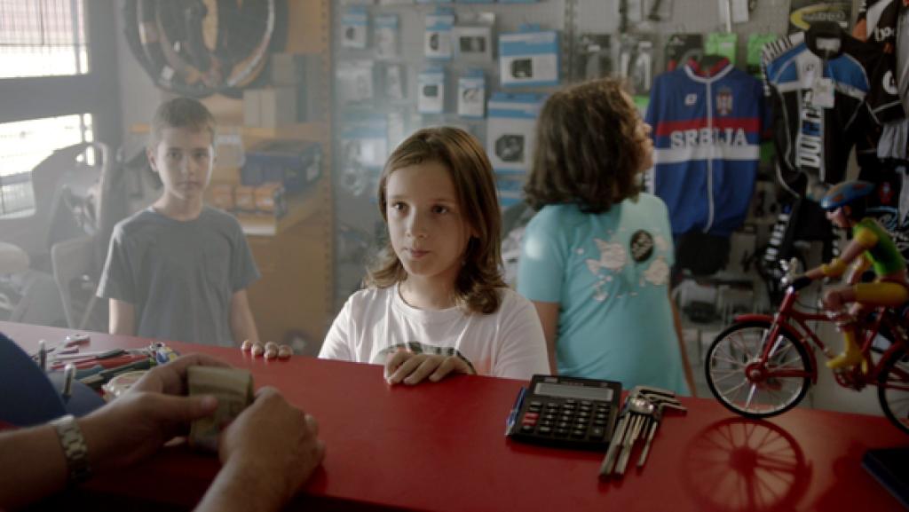 Ein Verkaufstresen in einem Fahrradgeschäft. Darauf links die Hände des Verkäufers, die einen Packen Geldscheine halten. Hinter dem Tresen ein Junge, der erwartungsvoll zum Verkäufer schaut. Im Hintergrund zwei Kinder, die sich im Laden umschauen.