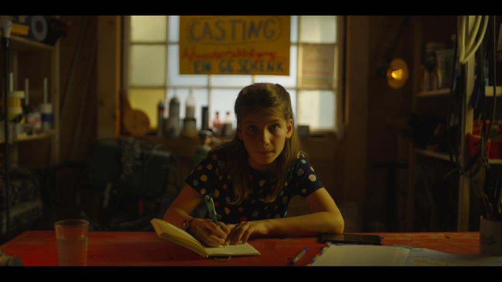 Rechte: KiKA/eitelsonnenschein GmbH Eileen sitzt an einem Schreibtisch, vor sich ein Notizbuch. Sie hört einem Kind zu, das ihr gegenüber sitzt.