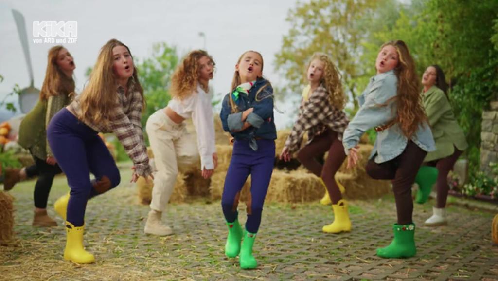 Kroni Pula und eine Gruppe von Mädchen tanzen in Gummistiefeln auf einem Hof.