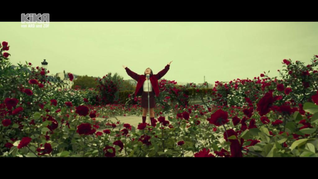 Lou hebt die Arme in die Luft, sie steht in einem Feld aus roten Rosen.