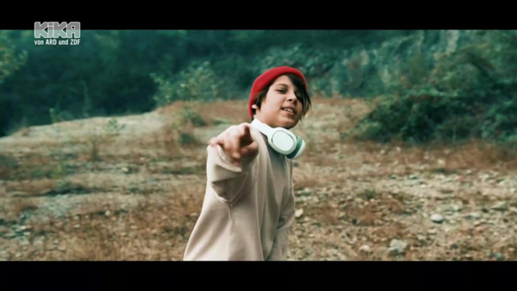 Leonardo zeigt in die Kamera. Er trägt eine Wollmütze und Kopfhörer um den Hals.