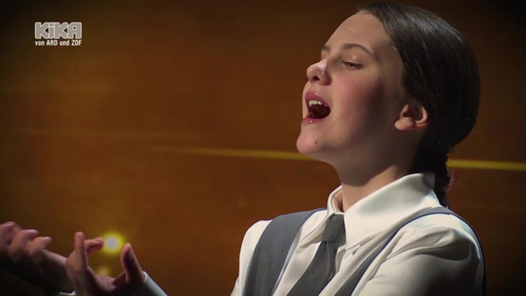 Asja Džogović singt. Sie trägt ein weißes Hemd und eine graue Krawatte.