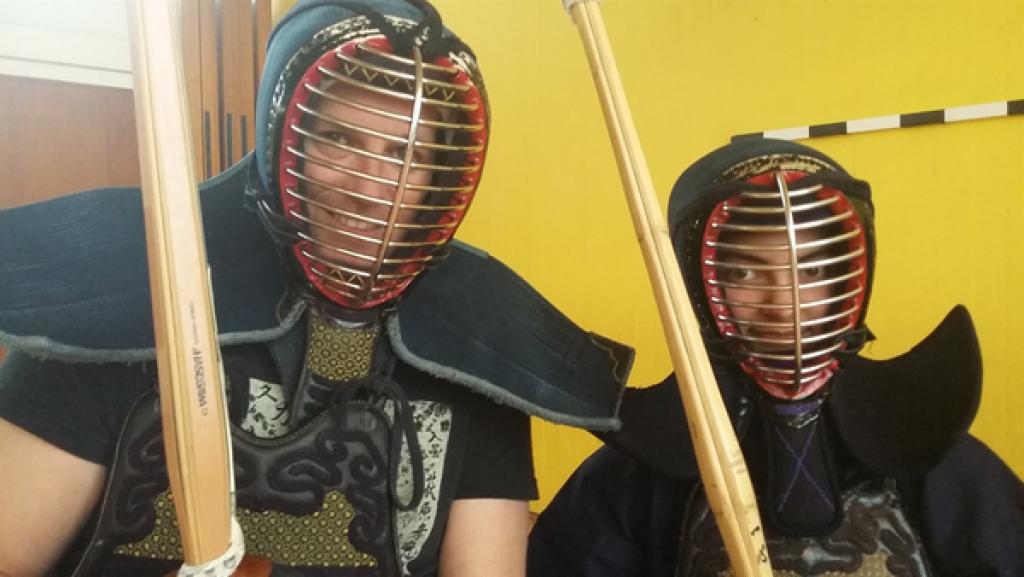 Rechte: KiKA Ben und Sanjana in Kendo-Gewand und Schutzhelm. Sie halten jeweils ein Bambusschwert vor sich.