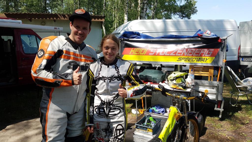 Rechte: KiKA/Anna Leistner Ben steht neben Speedway-Fahrerin Heidi in Motorradmontur. Hinter ihnen ein Plakat mit "Heidi Stolzenberg Speedway".