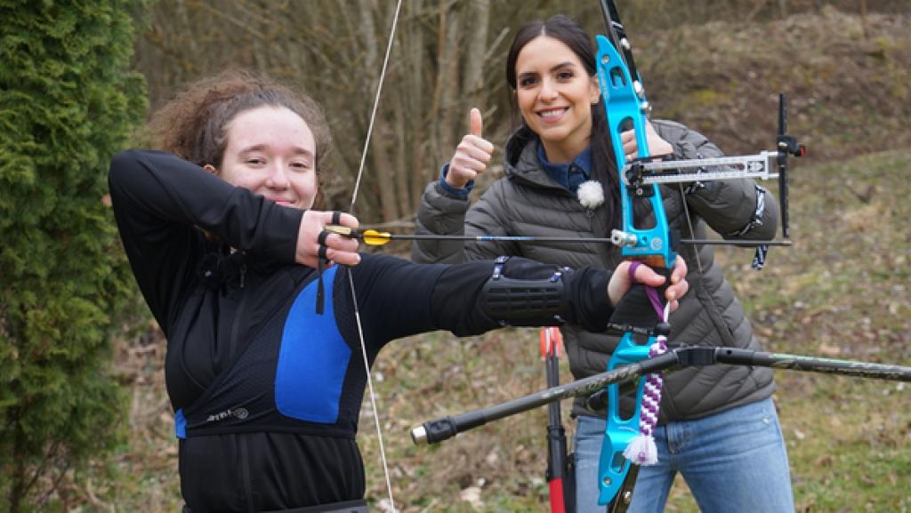 Rechte: KiKA/Franziska Gruber Vorne links steht Nevana, die in die Kamera lächelt. Dabei hält sie Bogen und Pfeil und hat eine Position zum Bogenschießen eingenommen. Dahiner steht Jess, die in die Kamera lächelt und Daumen hoch zeigt.