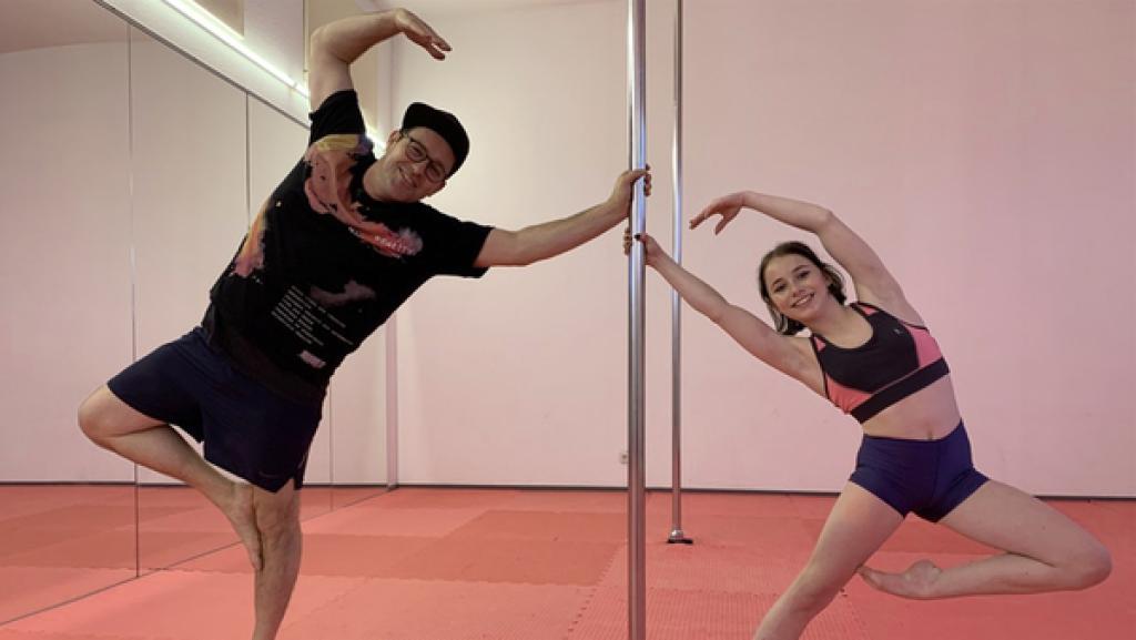 Rechte: KiKA/Eva Knäusl In einem Tanzssal ist in der Mitte eine Poledance-Stange. Links davon Ben, der auf einem Bein steht und sich an der Stange festhält. Rechts ist Finja, die ebenfalls auf einem Bein steht und sich mit einer Hand an der Stange festhält.