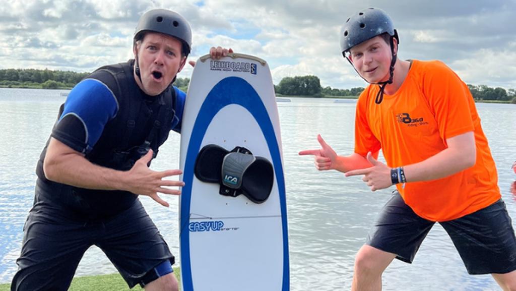 Rechte: KiKA/Jessica Schöne Links Ben, der einen Helm und ein Neoprenanzug trägt. Er stützt sich rechts auf ein Wakeboard. Rechts daneben steht Wakeboarder Fynn, der auf das Wakeboard in der Mitte zeigt.
