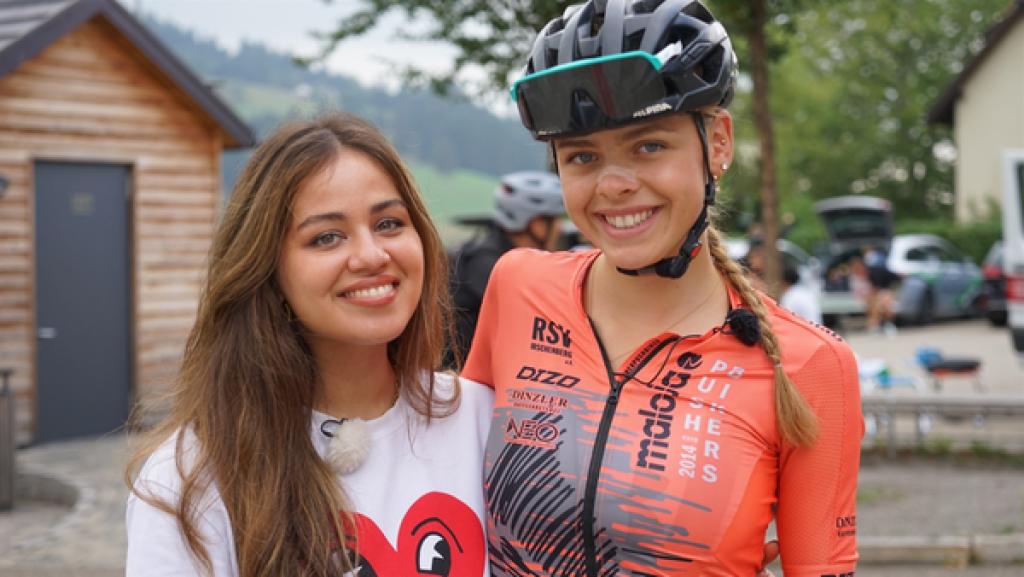 Rechte: KiKA/Franziska Gruber Sarah und Radsportlerin Antonia im Frontalfoto. Sie umgreifen sich für das Foto mit ihren Armen. Antonia ist in voller Radfahrerinnenmontur: Helm, Trikot, Brille auf dem Helm hochgezogen. Auf der Nase trägt Antonia ein horizontal gekleptes Pflaster.