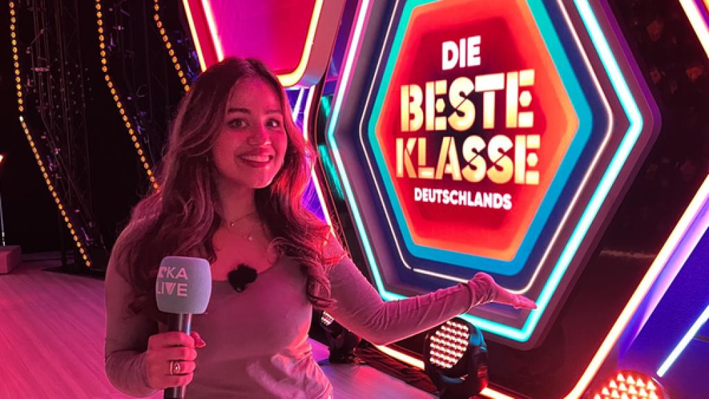 Rechte: KiKA/Oskar Lohaus Sarah mit einem 'KiKA LIVE' Mikrofon steht in pink getauchtem Licht vor einem leuchtenden Schild mit der Aufschrift "Die Beste Klasse Deutschlands".