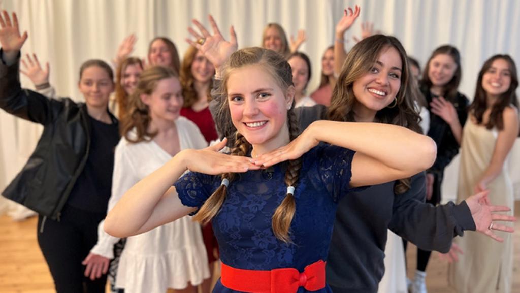 Rechte: KiKA/Jess Schöne Jeanne steht vorn und hält ihre Fingerspitzen aneinander unter ihr Gesicht. Hinter ihr steht Sarah. Im Hintergrund sind weitere Schülerinnen der Musicalschule zu sehen.