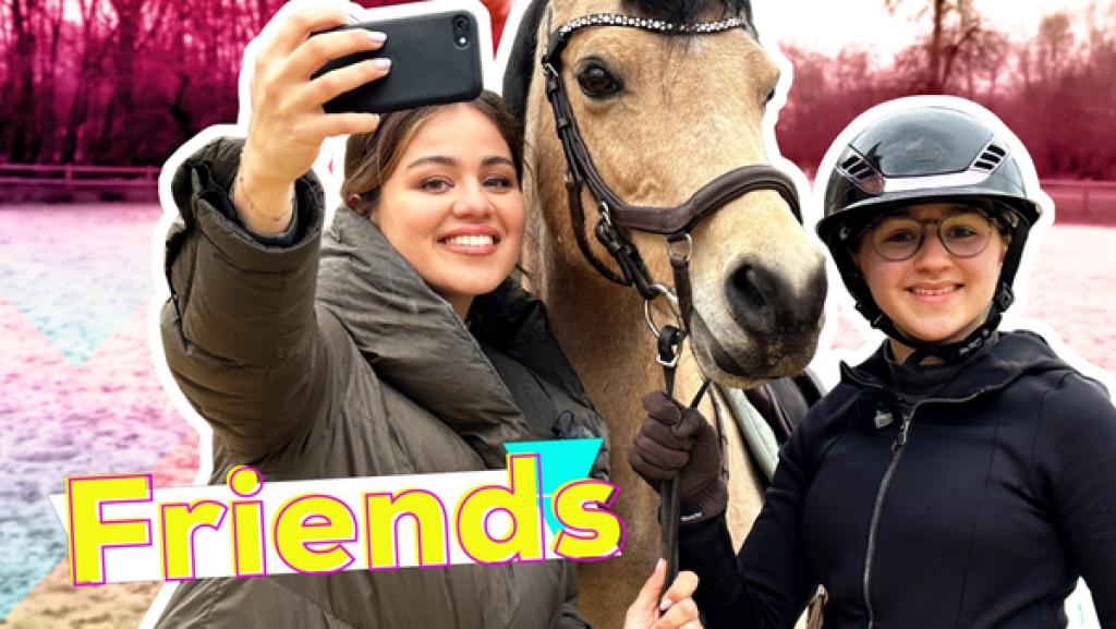 In der Mitte steht ein Pferd. Auf der einen Seite steht Lia daneben. Auf der anderen Seite steht Sarah, die ihren Arm ausstreckt und das Handy hoch hält.