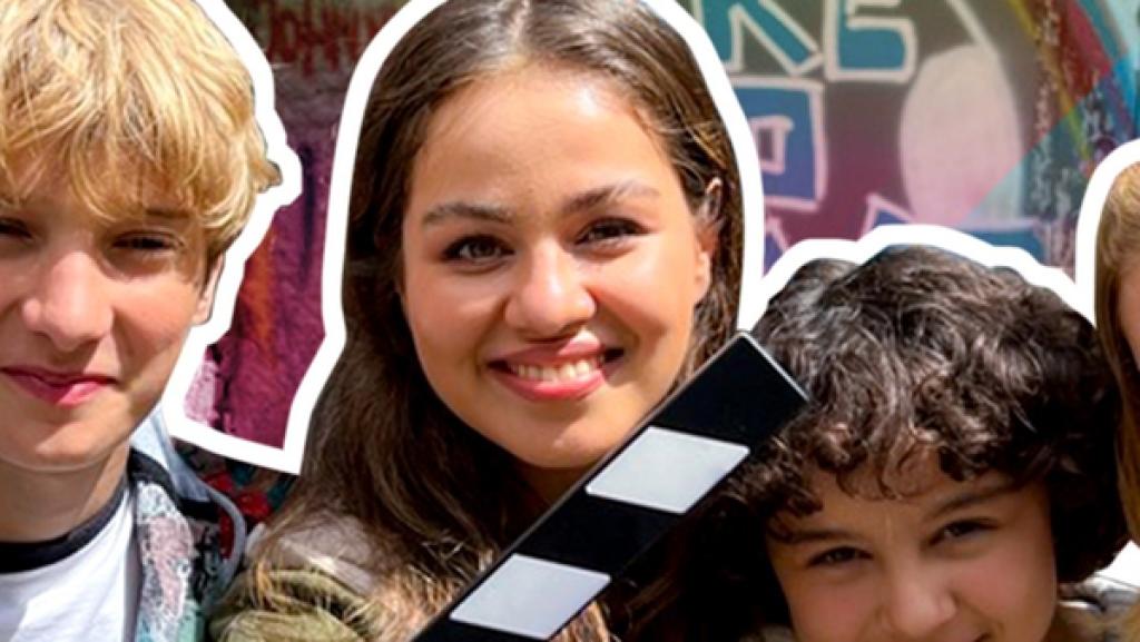 Moderatorin Sarah zusammen mit den Schloss Einstein-Darstellern Jonah, Janissa und Caroline. Vor ihnen eine Filmklappe und der pinke Schriftzug Am Set von Schloss Einstein. Sie lächeln in die Kamera. Hinter ihnen eine Wand mit Graffitti.