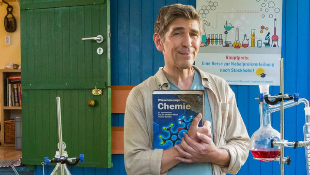 Fritz steht mit einer einem Chemiebuch an einem Tisch mit einem chemischen Versuchsaufbau - im Hintergrund der Bauwagen