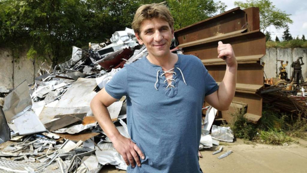 Fritz Fuchs zeigt auf einen hinter ihm liegenden Schrotthaufen. Eine Frau in lilafarbener Kleidung steht links von ihm.