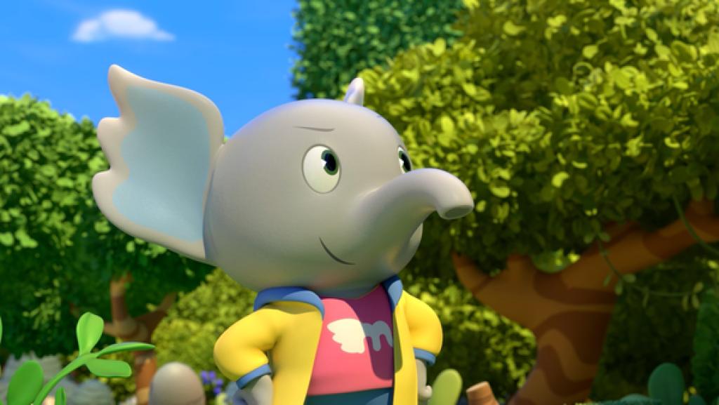 Elefant Mamfie steht vor Bäumen. Er schaut sich suchend um und lächelt.