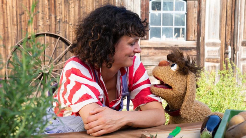 Die Landwirtin Saida vom Leckerhof und Moppi stehen zusammen hinter einem Tisch und schauen sich an. Vor ihnen auf dem Tisch befinden sich Kartoffeln, Tomaten, eine Aubergine und ein Kräutertopf. Sie befinden sich draußen, im Hintergrund ist ein Stallgebäude zu sehen.