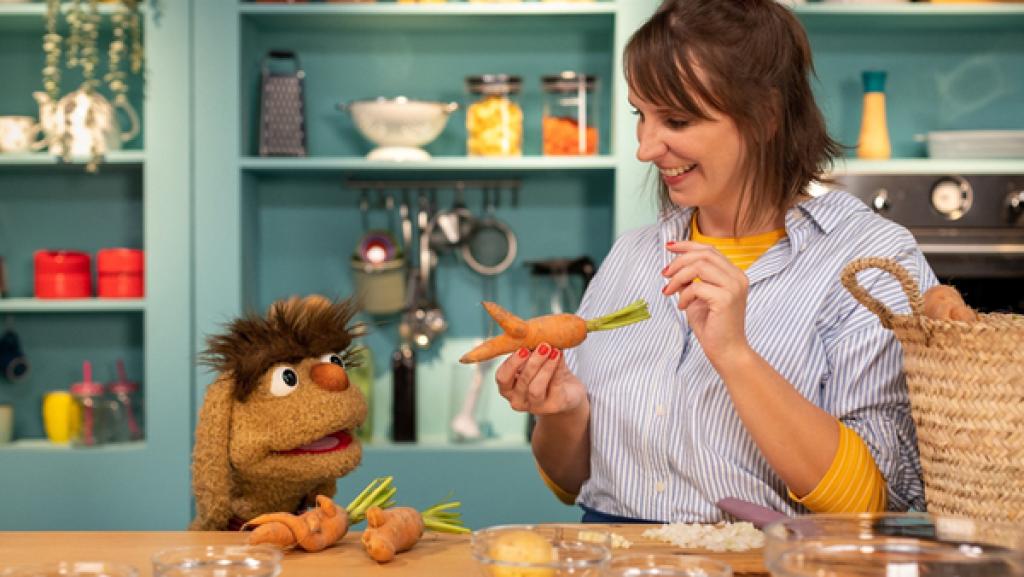 Moppi und Maya stehen in der Leckerküche an der Arbeitsplatte. Maya hält eine ungewöhlich geformte Karotte in der Hand. Sie erinnert an eine Maus. Die beiden staunen über die Karotte. Vor ihnen stehen Schüsseln mit Lebensmitteln, die für die Linsensuppe benötigt werden. Im Hintergrund befinden sich Regale mit Küchenutensilien und Lebensmitteln.