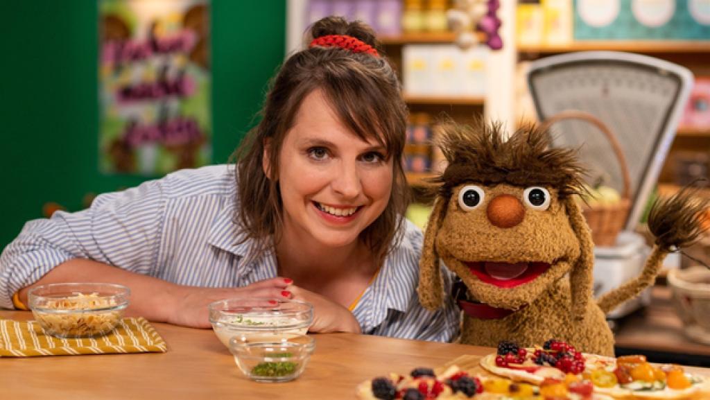 Maya und Moppi strahlen in die Kamera. Vor ihnen auf dem Tresen steht ein Brett mit "Doppimoppis". Diese Flammkuchen sind bunt belegt: auf der einen Hälfte verschiedenes Gemüse und auf der anderen Hälfte verschiedene Beeren. Auf dem Tresen befinden sich noch drei Schüsseln mit Belag-Zutaten. Im Hintergrund ist der Leckerladen mit seinen Lebensmittelregalen.