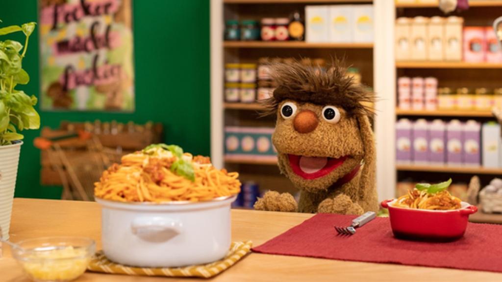 Moppi steht am Tresen im Leckerladen. Er schaut gierig auf einen großen Topf voll Spaghetti. Neben dem Topf steht unter anderem ein Basilikum, eine Flasche Öl, eine Salz- und eine Pfeffermühle. Im Hintergrund befinden sich die mit Lebensmitteln gefüllten Regale des Leckerladens.