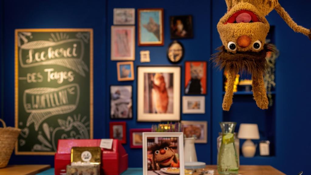 Moppis Kopf ist oben rechts im Bild zu sehen. Er scheint kopfüber von der Decke zu hängen, seine Ohren baumeln nach unten. Unter ihm steht auf dem Tresen ein Teller voller Wuffeln - herzförmige Waffeln mit einer Mangoldfüllung. Auf dem Tresen befindet sich außerdem die Kasse des Leckerladens, ein gerahmtes Bild von Moppi und eine Karaffe voller Wasser. Im Hintergrund hängen gerahmte Katzenbilder an einer Wand. An einer Tafel steht das Gericht des Tages: Wuffeln.