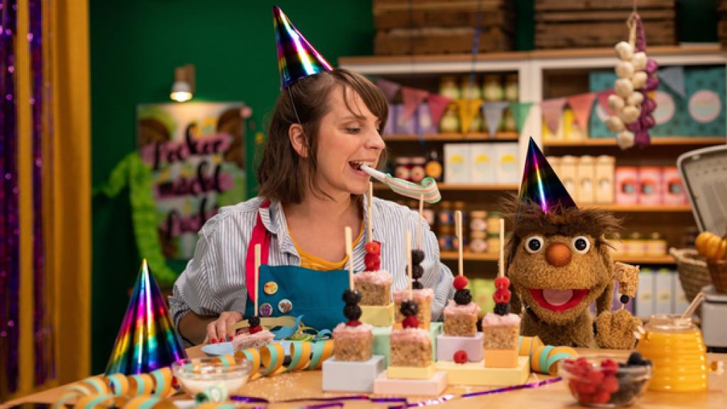 Maya und Moppi tragen bunte Partyhütchen. Maya hat eine Papiertröte im Mund und schaut zu Moppi. Moppi schaut vorfreudig in Richtung Partykuchen am Stiel vor ihm auf dem Tresen. Die Kuchen sind viereckig mit einem Holzstiel, auf dem Himbeeren und Brombeeren aufgespießt sind. Auf dem Tresen weitere Partydeko, eine Schale mit Beeren und ein Glas Honig. Im Hintergrund die mit Lebensmitteln gefüllten Regale des Leckerladens.