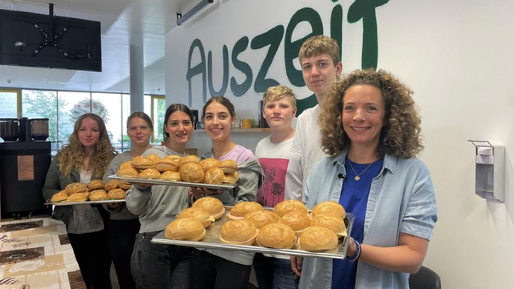 Gesa steht mit Schüler:innen im Café Auszeit. Sie halten Tablette mit geschmierten Brötchen in der Hand.