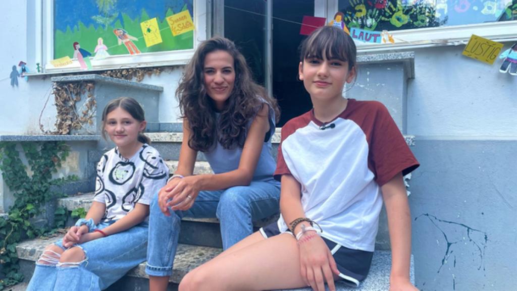 Mona sitzt zwischen Leijla (11) und Martha (10) auf den Treppen am Eingang eines Jugendzentrums.