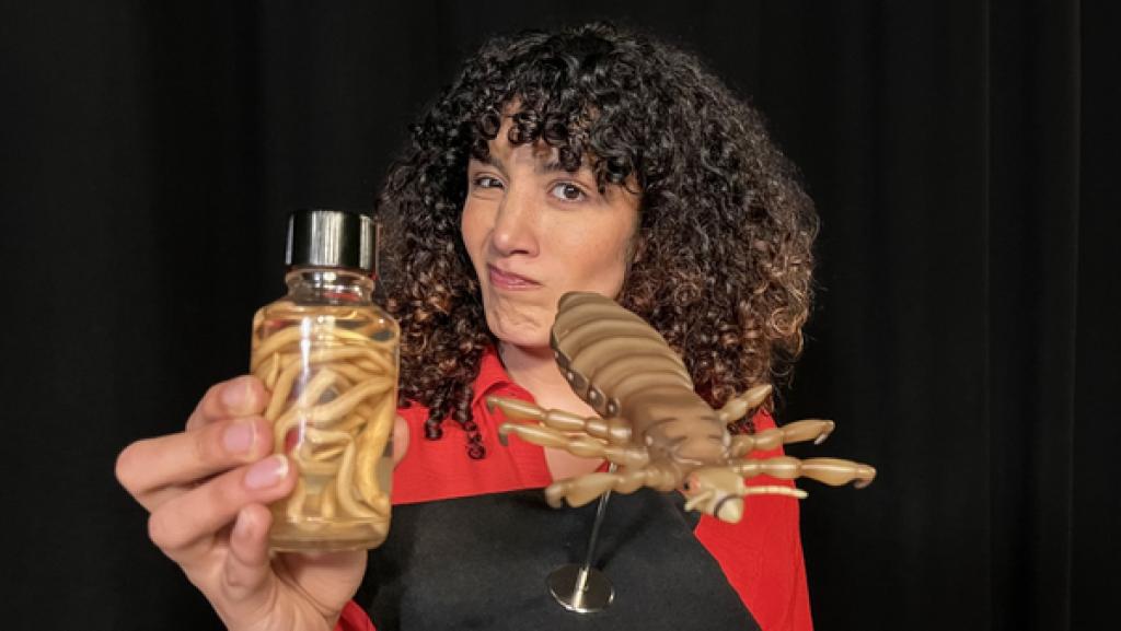 Reporterin Tessniem hält ein Glas mit einem Spulwurm und das Modell einer Kopflaus in die Kamera.