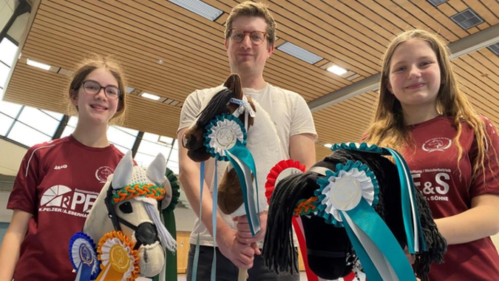 Robert steht zwischen Rebecca und Melina in einer Turnhalle. Alle drei halten ein Steckenpferd in ihren Händen.