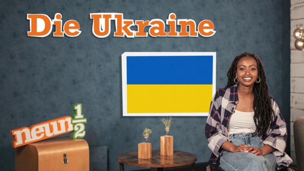Luam sitzt auf einem Stuhl neben einem kleinen Tisch und dem neuneinhalb-Logo. Auf einer blauen Wand steht der Schriftzug "Die Ukraine" und hinter Luam ist die ukrainische Flagge zu sehen.