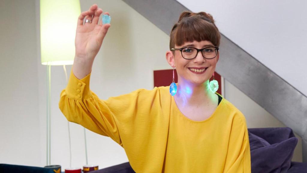 Princess of Science - Folge 16 - Kluge Kleidung | Rechte: Bojan Ritan Moderatorin Patrizia trägt grün und blau leuchtende Stein-Ohrringe und hält einen weiteren in der Hand nach oben. Dabei lächelt sie in die Kamera.